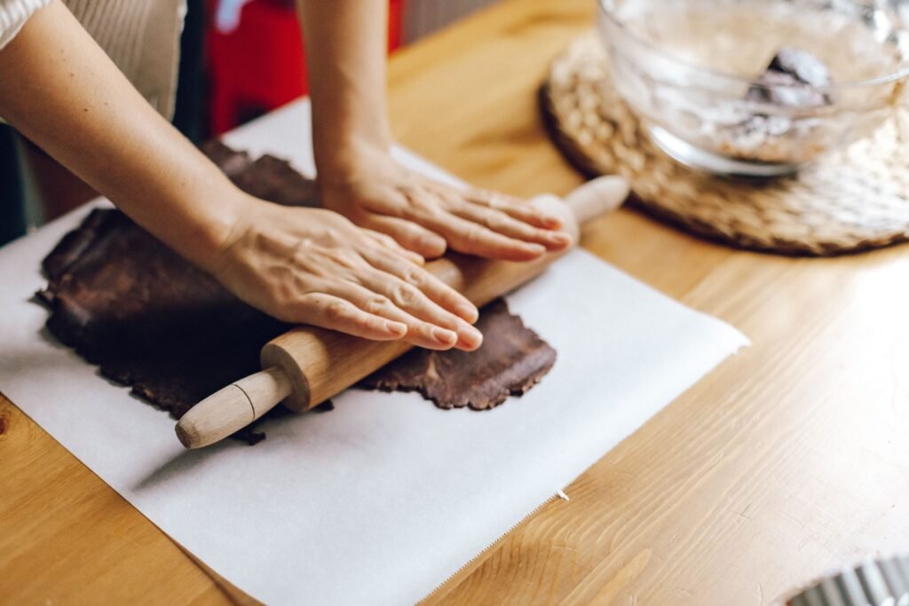 Hands rolling chocolate dough with a rolling pin on parchment in a home kitchen setting.